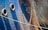 A close-up view of the hull of a ship displaying two large tires hanging on the side, likely used as fenders. Multiple heavy chains are draped across the side, casting shadows against the metal surface. The dark blue metallic hull shows some signs of rust and weathering, contributing to a rugged, industrial texture.