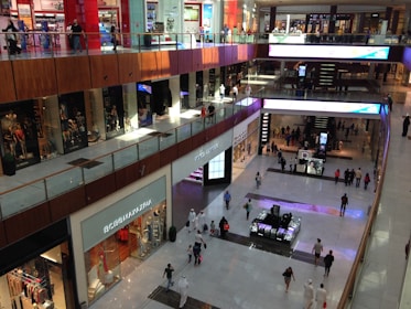 A multi-level shopping mall with various storefronts illuminated by bright lights. People are seen walking on both levels, some visiting the stores. The architecture features modern design with glass railings and polished floors.