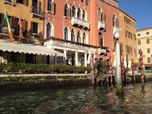A picturesque scene of a Venetian canal with a historic building featuring ornate balconies and arched windows. The façade is painted in warm, earthy tones and is adorned with multiple flags. Decorative poles with red and white stripes line the water's edge, reflecting in the calm canal.