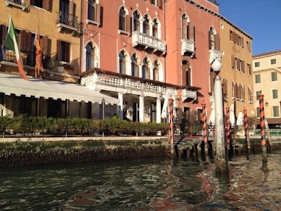 A picturesque scene of a Venetian canal with a historic building featuring ornate balconies and arched windows. The façade is painted in warm, earthy tones and is adorned with multiple flags. Decorative poles with red and white stripes line the water's edge, reflecting in the calm canal.