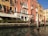 A picturesque scene of a Venetian canal with a historic building featuring ornate balconies and arched windows. The façade is painted in warm, earthy tones and is adorned with multiple flags. Decorative poles with red and white stripes line the water's edge, reflecting in the calm canal.