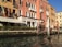 A picturesque scene of a Venetian canal with a historic building featuring ornate balconies and arched windows. The façade is painted in warm, earthy tones and is adorned with multiple flags. Decorative poles with red and white stripes line the water's edge, reflecting in the calm canal.