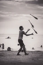 A person is juggling clubs on a beach during what appears to be a cloudy day. The background features people swimming and playing in the water, with a few small boats on the horizon. The scene is set in a grayscale tone, capturing a peaceful, yet dynamic moment.
