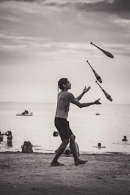 A person is juggling clubs on a beach during what appears to be a cloudy day. The background features people swimming and playing in the water, with a few small boats on the horizon. The scene is set in a grayscale tone, capturing a peaceful, yet dynamic moment.