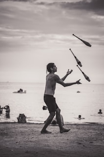 A person is juggling clubs on a beach during what appears to be a cloudy day. The background features people swimming and playing in the water, with a few small boats on the horizon. The scene is set in a grayscale tone, capturing a peaceful, yet dynamic moment.