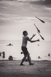 A person is juggling clubs on a beach during what appears to be a cloudy day. The background features people swimming and playing in the water, with a few small boats on the horizon. The scene is set in a grayscale tone, capturing a peaceful, yet dynamic moment.