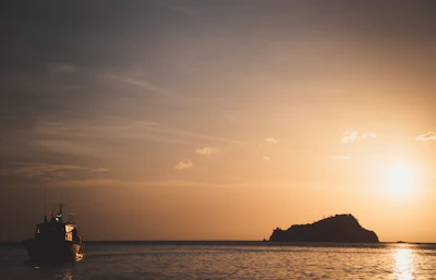 Sunset view over the ocean with silhouettes of the boat and island, glowing in warm tropical hues.