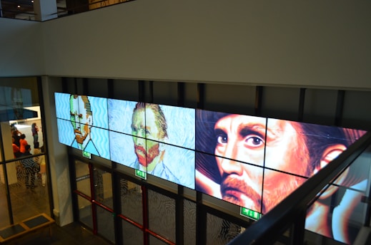 A series of three digital panels featuring artwork resembling portraits displayed along a wall in an interior setting. The portraits vary in artistic style, transitioning from pixel art to post-impressionism, each depicting a face with different expressions. Below the panels, glass doors and emergency exit signs are visible. A group of people appears in the background on the left side.