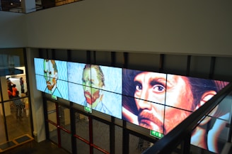 A series of three digital panels featuring artwork resembling portraits displayed along a wall in an interior setting. The portraits vary in artistic style, transitioning from pixel art to post-impressionism, each depicting a face with different expressions. Below the panels, glass doors and emergency exit signs are visible. A group of people appears in the background on the left side.