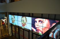 A series of three digital panels featuring artwork resembling portraits displayed along a wall in an interior setting. The portraits vary in artistic style, transitioning from pixel art to post-impressionism, each depicting a face with different expressions. Below the panels, glass doors and emergency exit signs are visible. A group of people appears in the background on the left side.
