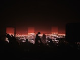 A concert stage with a silhouette of musicians playing instruments and performing. The background features a vibrant cityscape illuminated with bright lights during nighttime. Some audience members' hands and heads are visible in the foreground, adding to the energetic concert atmosphere.