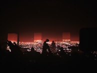 A concert stage with a silhouette of musicians playing instruments and performing. The background features a vibrant cityscape illuminated with bright lights during nighttime. Some audience members' hands and heads are visible in the foreground, adding to the energetic concert atmosphere.