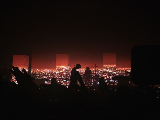 A concert stage with a silhouette of musicians playing instruments and performing. The background features a vibrant cityscape illuminated with bright lights during nighttime. Some audience members' hands and heads are visible in the foreground, adding to the energetic concert atmosphere.