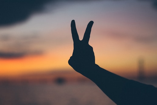 A silhouette of a hand making a peace sign is seen against the backdrop of a vibrant sunset, with a range of warm colors like orange and yellow blending into darker shades near the horizon.