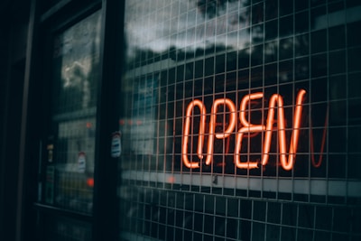 A glowing neon sign displaying the word 'OPEN' is affixed to a grid-like glass window. The sign emits a warm orange light, contrasting with the reflections and darker tones of the window and surrounding area, suggesting a moody or late evening setting.