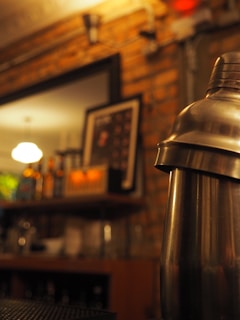 A dimly lit bar setting featuring a metallic cocktail shaker in focus. The background includes shelves with bottles and a blurred picture frame on a brick wall.