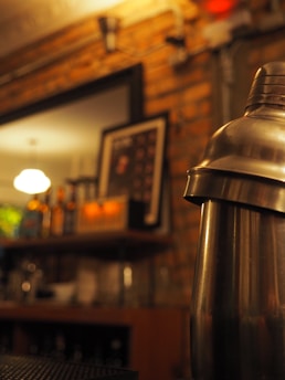 A dimly lit bar setting featuring a metallic cocktail shaker in focus. The background includes shelves with bottles and a blurred picture frame on a brick wall.