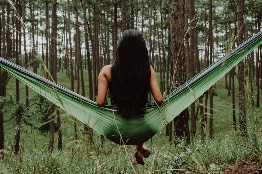 A woman with long hair sits on a green hammock facing a forest of trees