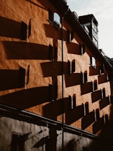 An architectural facade with warm, textured walls casting long shadows from numerous small windows. The lighting creates a dramatic effect with alternating light and shadow patterns, highlighting the architectural details.
