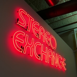 A neon sign displaying the words 'STEREO EXCHANGE' is mounted on a wall. The sign emits a bright red glow and is seen in a slightly dark room with a decorated ceiling and some piping visible above.