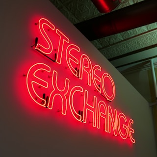 A neon sign displaying the words 'STEREO EXCHANGE' is mounted on a wall. The sign emits a bright red glow and is seen in a slightly dark room with a decorated ceiling and some piping visible above.