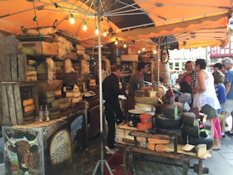 A bustling market stall featuring a variety of cheeses is displayed under a canopy. The cheeses are stacked on wooden crates and woven baskets, with small chalkboard signs indicating their types. Several people are engaged in conversation with the vendor, possibly tasting or purchasing the cheeses. The scene is lively and vibrant, with warm lighting from hanging bulbs creating an inviting atmosphere.