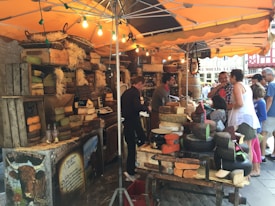 A bustling market stall featuring a variety of cheeses is displayed under a canopy. The cheeses are stacked on wooden crates and woven baskets, with small chalkboard signs indicating their types. Several people are engaged in conversation with the vendor, possibly tasting or purchasing the cheeses. The scene is lively and vibrant, with warm lighting from hanging bulbs creating an inviting atmosphere.
