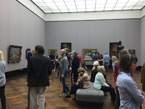 A group of people are gathered in an art gallery, observing various paintings displayed on the walls. The room has white walls and wooden flooring, with a modern, minimalistic ceiling design. Some individuals are listening to audio guides.