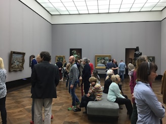 A group of people are gathered in an art gallery, observing various paintings displayed on the walls. The room has white walls and wooden flooring, with a modern, minimalistic ceiling design. Some individuals are listening to audio guides.