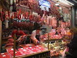 A bustling deli or meat store featuring a wide variety of hanging cured meats like sausages and hams. Two workers behind the counter carve meat on a slicer, while a customer at the counter holds a shopping bag. The counter displays various cold cuts and other deli items, with numerous packaged goods stacked on shelves.