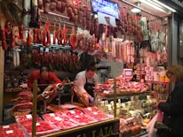 A bustling deli or meat store featuring a wide variety of hanging cured meats like sausages and hams. Two workers behind the counter carve meat on a slicer, while a customer at the counter holds a shopping bag. The counter displays various cold cuts and other deli items, with numerous packaged goods stacked on shelves.