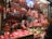 A bustling deli or meat store featuring a wide variety of hanging cured meats like sausages and hams. Two workers behind the counter carve meat on a slicer, while a customer at the counter holds a shopping bag. The counter displays various cold cuts and other deli items, with numerous packaged goods stacked on shelves.