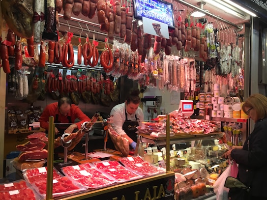 A bustling deli or meat store featuring a wide variety of hanging cured meats like sausages and hams. Two workers behind the counter carve meat on a slicer, while a customer at the counter holds a shopping bag. The counter displays various cold cuts and other deli items, with numerous packaged goods stacked on shelves.