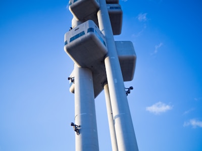Three large, modernist-style concrete columns rise into the clear blue sky. Along the columns, there are several dark, abstract sculptures resembling crawling figures. Each column features cuboid structures near the top, with tinted windows that add a futuristic look.