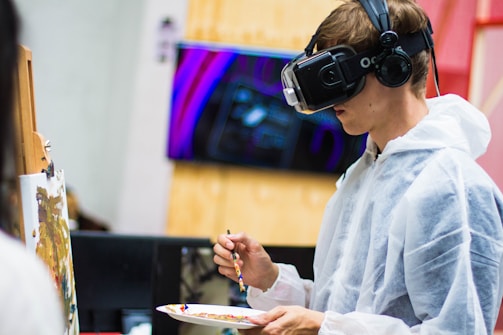 A person wearing a virtual reality headset and headphones is painting on a canvas. The individual is dressed in a white protective suit and holds a paintbrush and palette with various colors. In the background, there is a blurred image of digital art displayed on a screen.