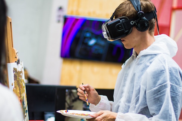 A person wearing a virtual reality headset and headphones is painting on a canvas. The individual is dressed in a white protective suit and holds a paintbrush and palette with various colors. In the background, there is a blurred image of digital art displayed on a screen.