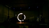 A performer is on stage, creating a mesmerizing circle of light with a tool in motion. The setting is dimly lit, emphasizing the glowing circle, while the performer appears as a silhouette against the dark backdrop.