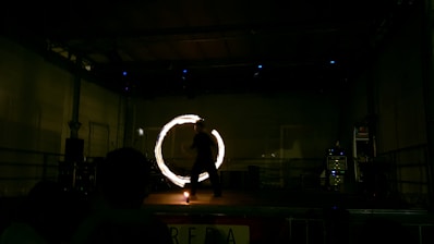 A performer is on stage, creating a mesmerizing circle of light with a tool in motion. The setting is dimly lit, emphasizing the glowing circle, while the performer appears as a silhouette against the dark backdrop.