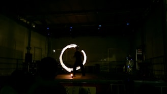 A performer is on stage, creating a mesmerizing circle of light with a tool in motion. The setting is dimly lit, emphasizing the glowing circle, while the performer appears as a silhouette against the dark backdrop.