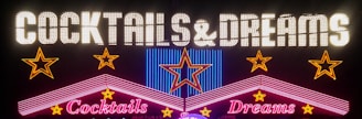 A vibrant neon sign featuring the words 'Cocktails & Dreams' with colorful stars and geometric shapes around it. The words are illuminated in bright neon lights, creating a lively and inviting atmosphere.