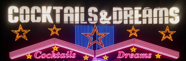 A vibrant neon sign featuring the words 'Cocktails & Dreams' with colorful stars and geometric shapes around it. The words are illuminated in bright neon lights, creating a lively and inviting atmosphere.