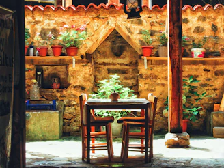 A rustic courtyard features a wooden table with two chairs, surrounded by a stone wall adorned with potted plants. Sunlight casts shadows on the stone pavement, creating a warm, inviting ambiance. A lantern hangs above the table, adding a charming touch.