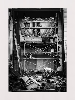A black and white photo of a construction site with scaffolding in front of a brick wall. A person is kneeling on the ground, surrounded by bricks and construction materials. Ladders are positioned on the sides, and various tools and equipment are scattered around.