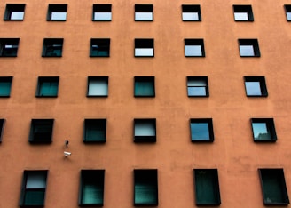 A building facade with a grid-like pattern of square windows against an orange-brown wall. The windows are dark and reflective, some partially open, adding a sense of symmetry and uniformity. A security camera is positioned among the windows.