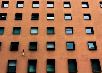A building facade with a grid-like pattern of square windows against an orange-brown wall. The windows are dark and reflective, some partially open, adding a sense of symmetry and uniformity. A security camera is positioned among the windows.