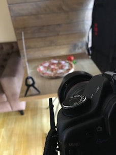 A camera is positioned on a tripod in front of a wooden table, focusing on a blurred pizza. The setup seems to be for a photo or video shoot. The background includes a soft chair and a wooden wall, with a red item possibly a tomato near the pizza.