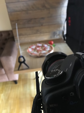 A camera is positioned on a tripod in front of a wooden table, focusing on a blurred pizza. The setup seems to be for a photo or video shoot. The background includes a soft chair and a wooden wall, with a red item possibly a tomato near the pizza.