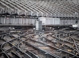 An intricate network of railway tracks converges towards a station surrounded by a high, curved metal roof with vertical supports. The tracks are equipped with various signal and electrical equipment. The area appears industrial, showcasing complex rail infrastructure.