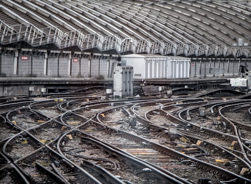 An intricate network of railway tracks converges towards a station surrounded by a high, curved metal roof with vertical supports. The tracks are equipped with various signal and electrical equipment. The area appears industrial, showcasing complex rail infrastructure.