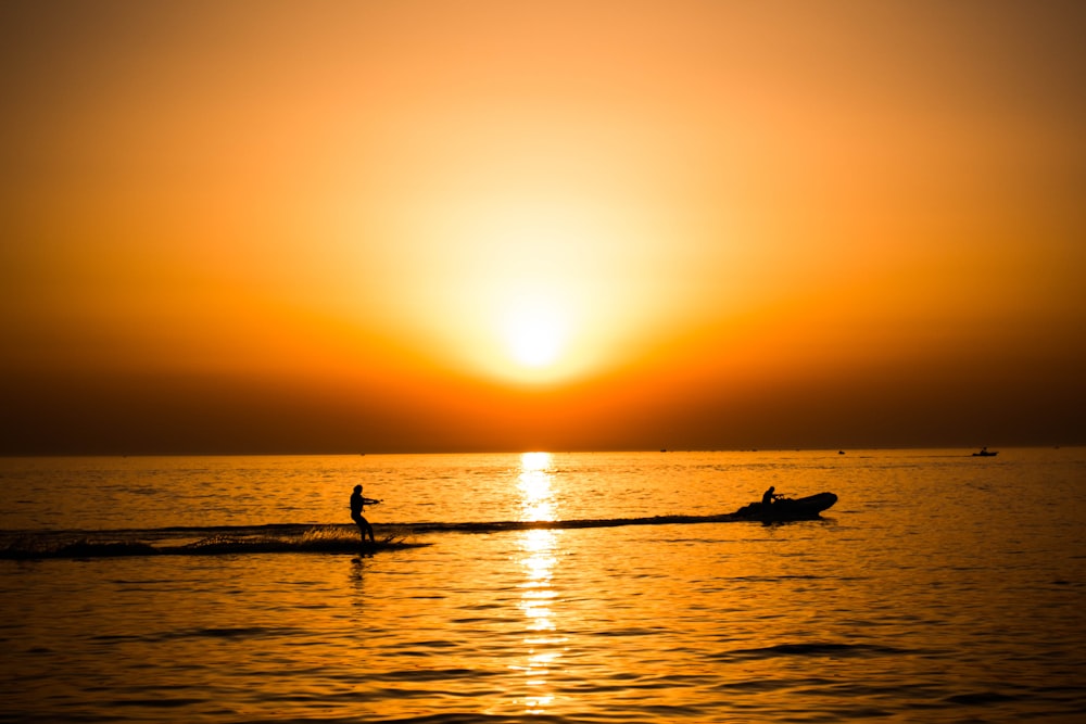 silhouette photo of man riding a motorboat with man surfboarding behind during golden hour